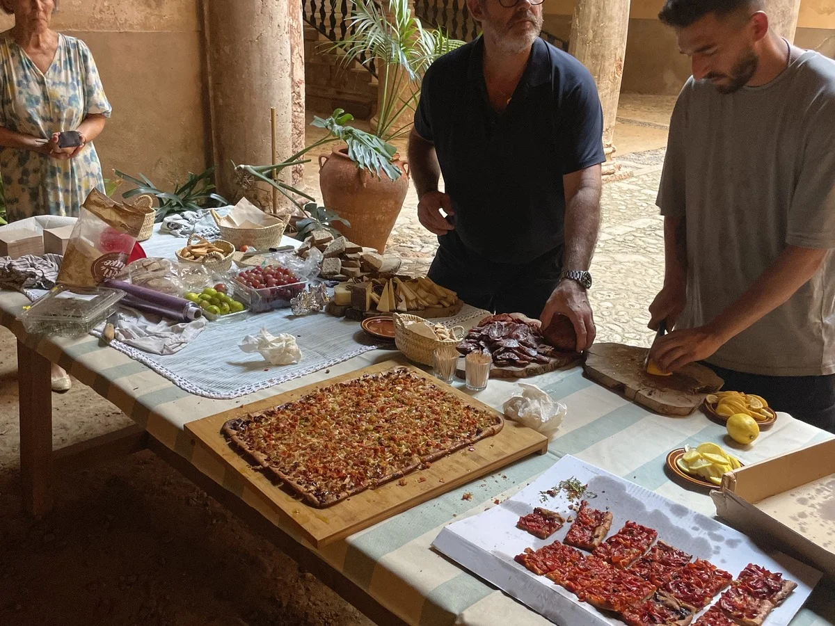 Personas preparando y compartiendo platos locales en una mesa larga dentro de un patio mediterráneo histórico, capturando una experiencia gastronómica auténtica en las Islas Baleares.