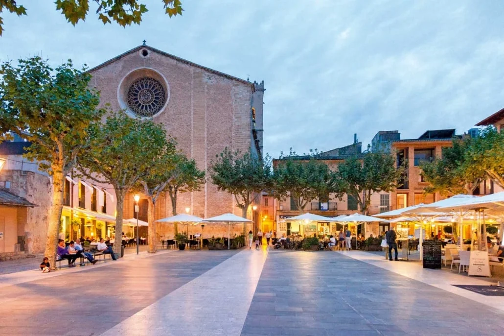 Plaça Major de Pollença al atardecer con terrazas de restaurantes y la Iglesia de Nuestra Señora de los Ángeles en Mallorca