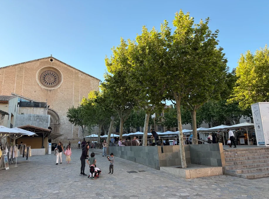 Plaça Major in Pollença, Mallorca during the day with outdoor cafés, local visitors, and historic architecture.