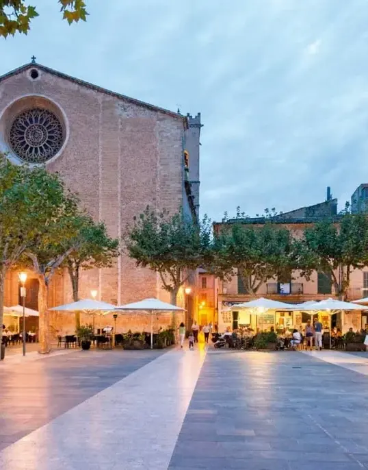 Plaça Major in Pollença at sunset with restaurant terraces and the Church of Our Lady of the Angels in Mallorca